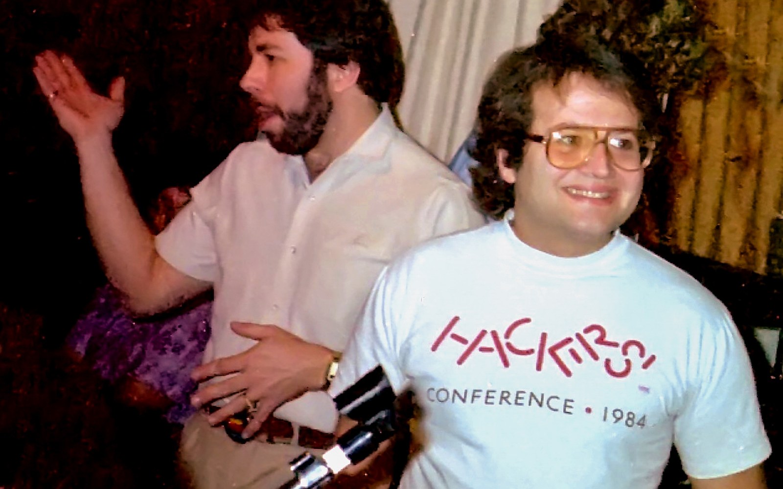 Two men at a gathering, one wearing tinted glasses and a Hackers Conference 1984 t-shirt