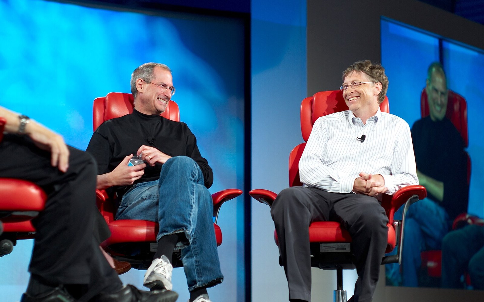 Two men laugh together while seated in red chairs during an on-stage conference interview