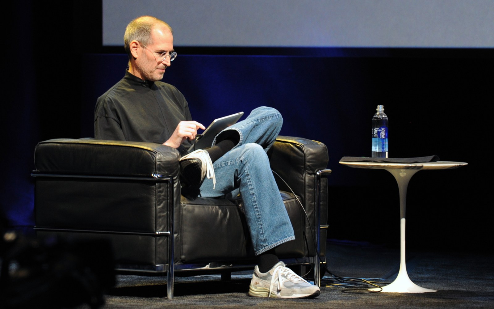 Man in black turtleneck sits in leather chair, tapping a tablet on stage