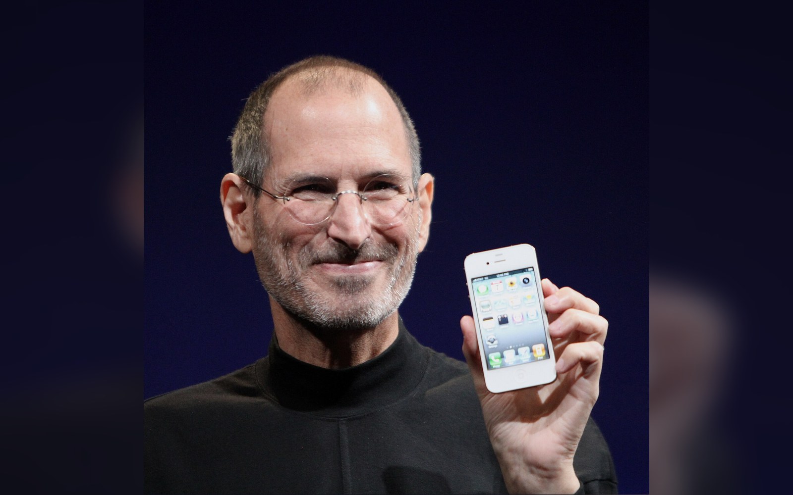 Man in glasses and black turtleneck holding up a white smartphone