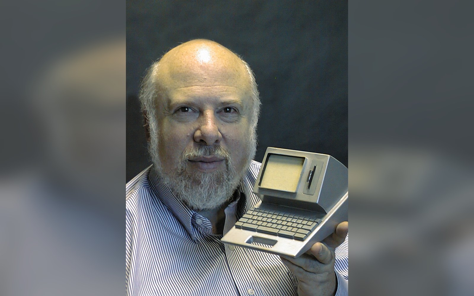 A bearded man holds up a small vintage portable computer with built-in keyboard and screen