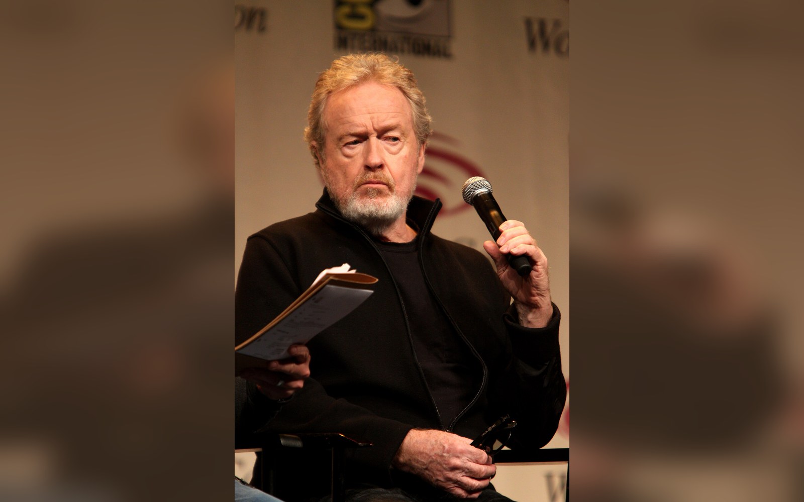 Ridley Scott holds a microphone and papers while speaking on a convention panel