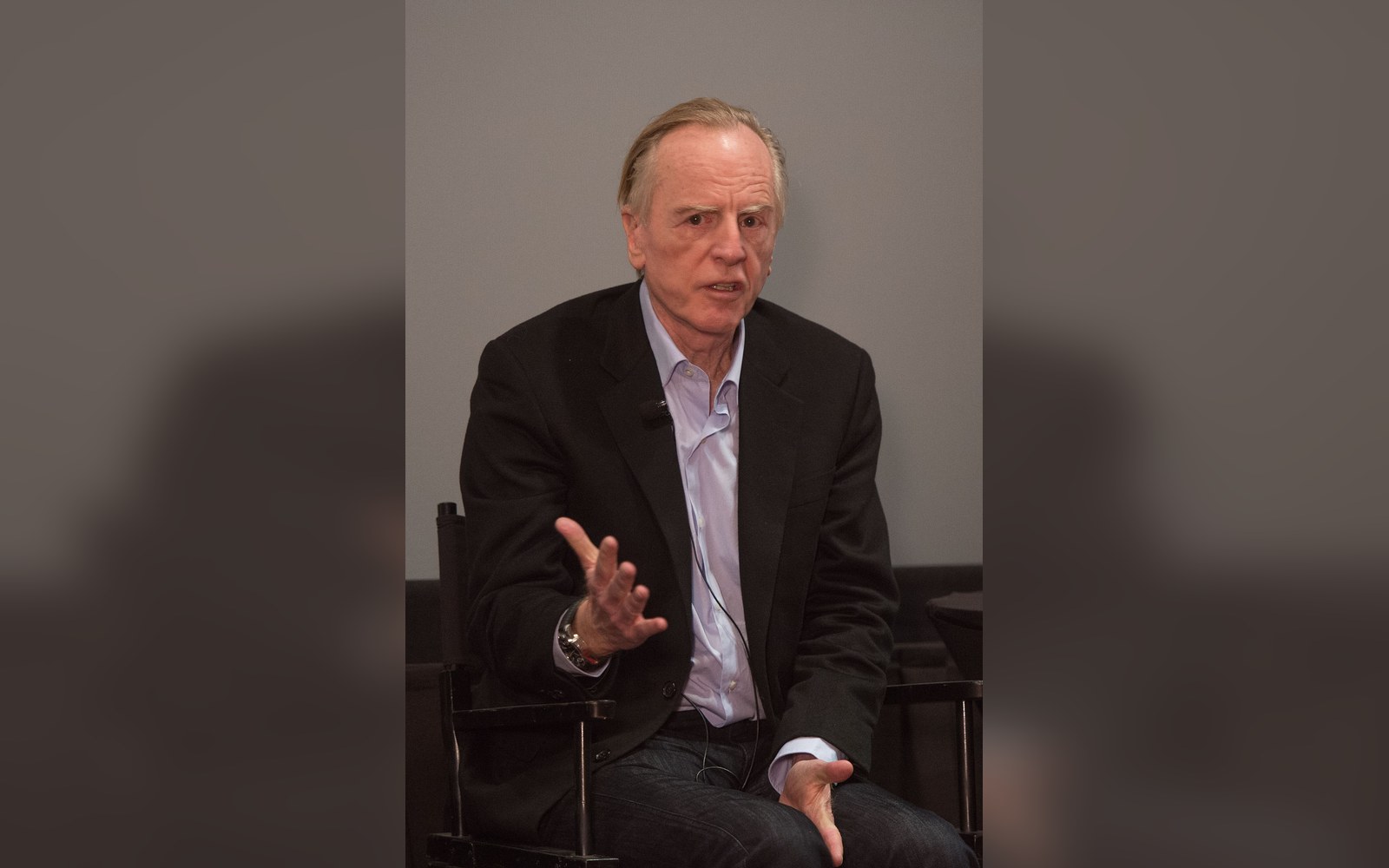 An older man in a blazer gestures with his hand while seated during an interview