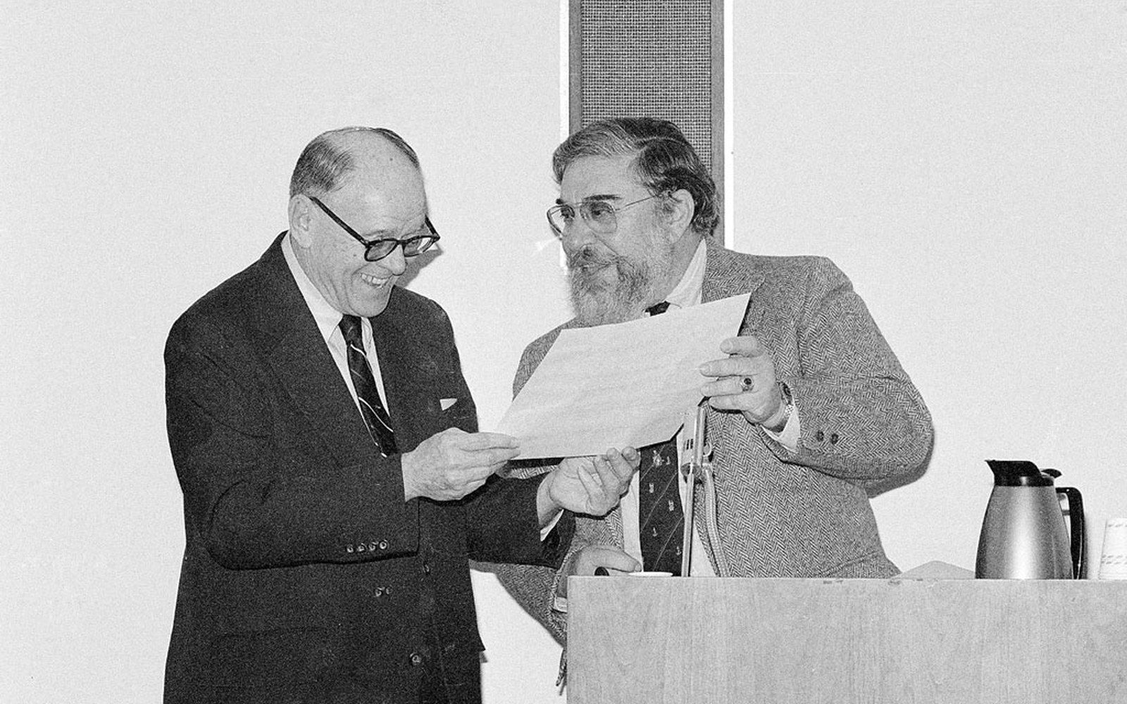 Two men in suits exchange a document at a podium during an award presentation