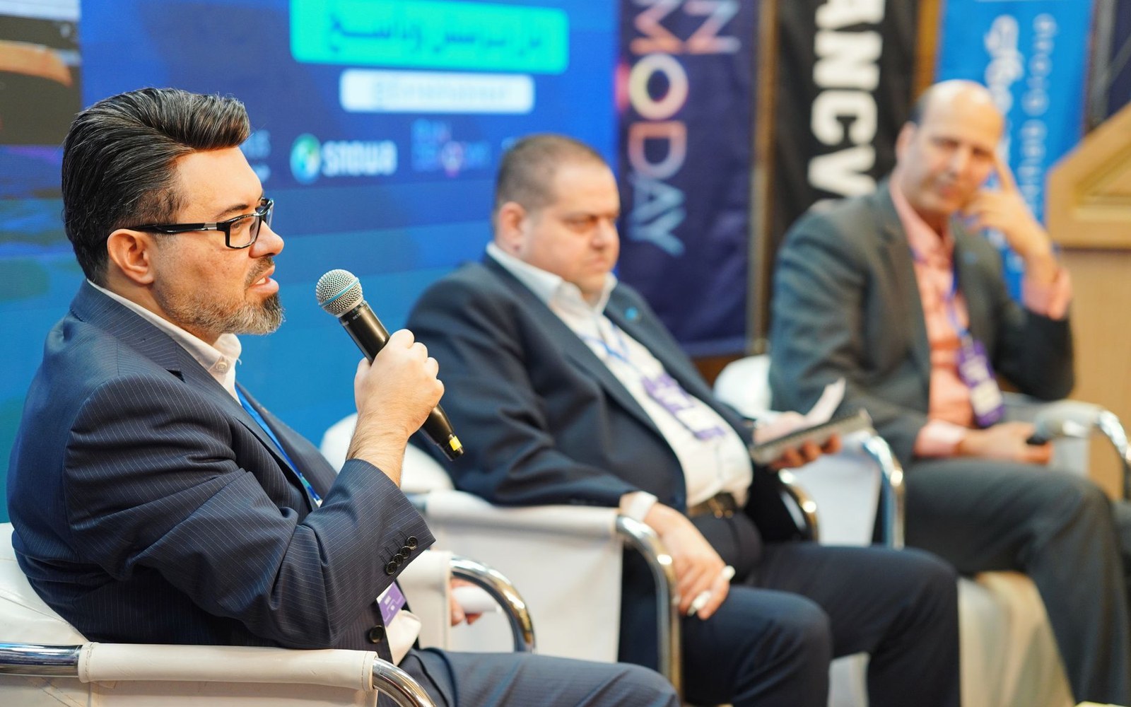 Man speaks into a microphone while seated on a panel with two other attendees