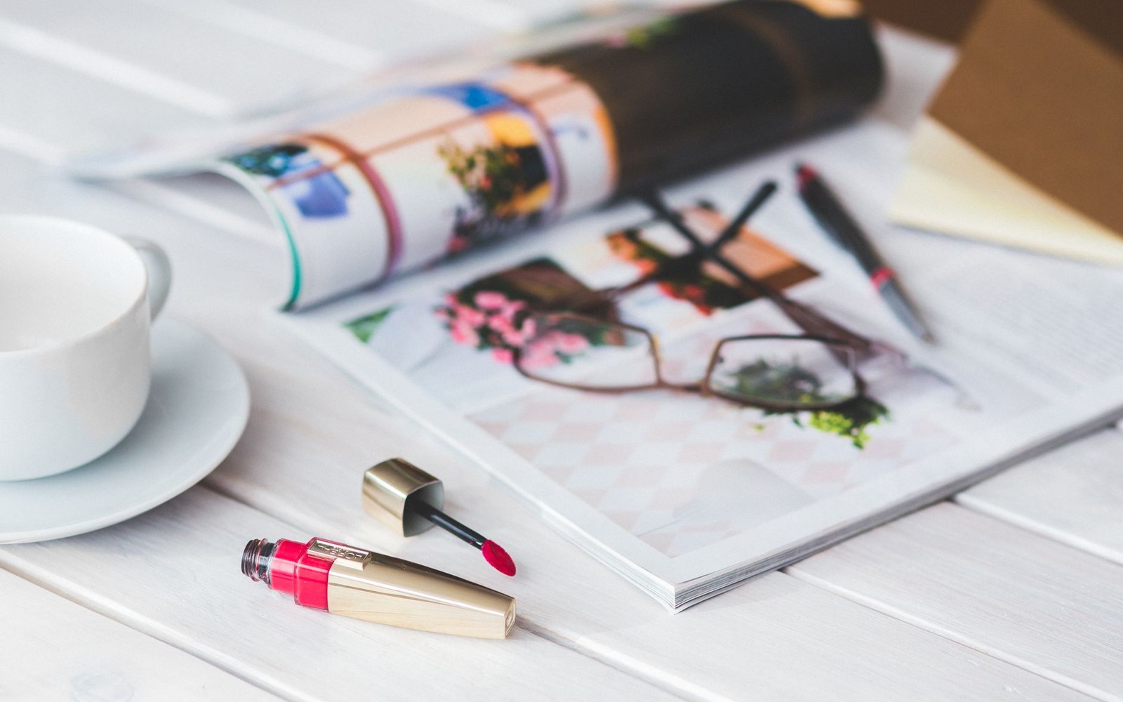 Open magazine, teacup and saucer, and a tube of pink lip gloss on a white table